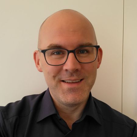 Peter Krautzberger, in front of a white, wooden door wearing a dark blue shirt and black glasses.